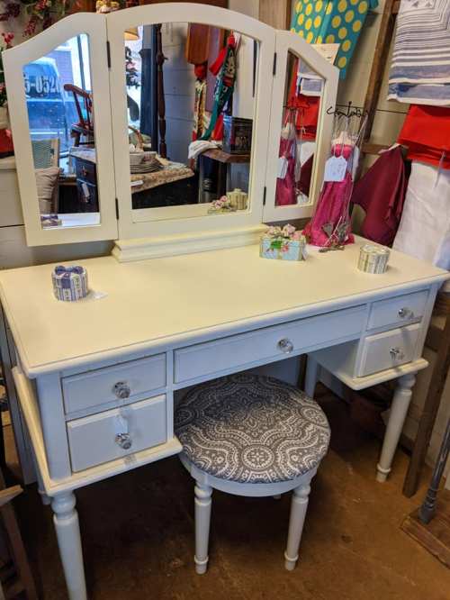 A white vanity with a mirror and three drawers, accompanied by a round, patterned stool.