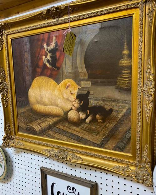 A framed painting of a yellow cat and a black kitten playing together on a rug, with a small ball between them.