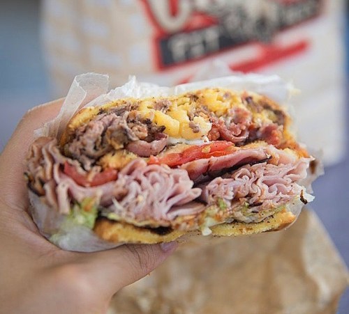 A hand holding a large, layered sandwich filled with various meats, cheese, lettuce, and tomato, wrapped in paper.