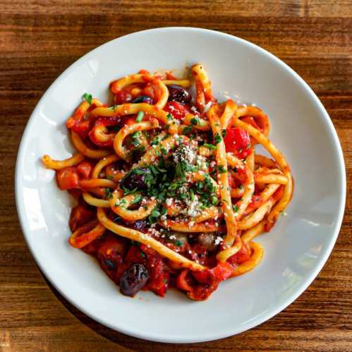 A bowl of pasta with tomato sauce, olives, and herbs, garnished with grated cheese on a wooden table.