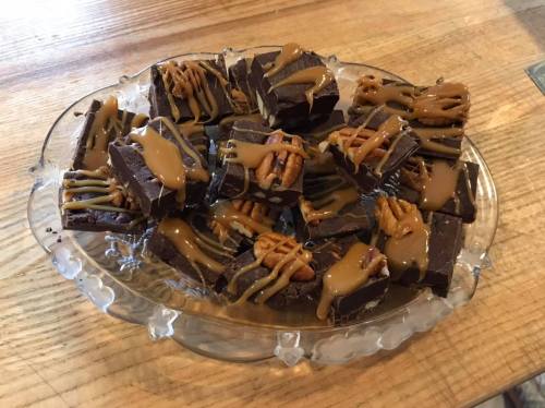 A glass platter filled with chocolate fudge squares drizzled with caramel and topped with pecans.