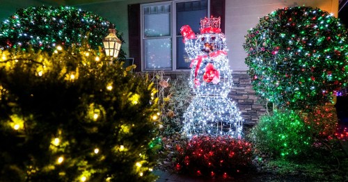 A brightly lit snowman decoration surrounded by colorful holiday lights and greenery in a festive outdoor setting.