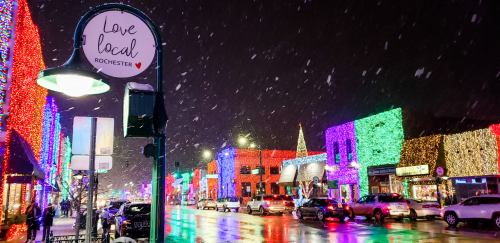 Colorful holiday lights illuminate a snowy street in Rochester, with a "Love Local" sign visible.