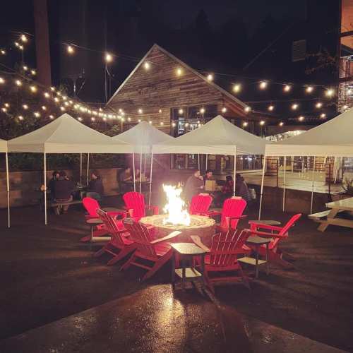 A cozy outdoor space at night with red chairs around a fire pit, surrounded by tents and string lights.