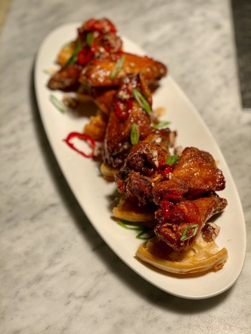 A platter of crispy chicken wings topped with red chili and green onions, served on a bed of waffles.