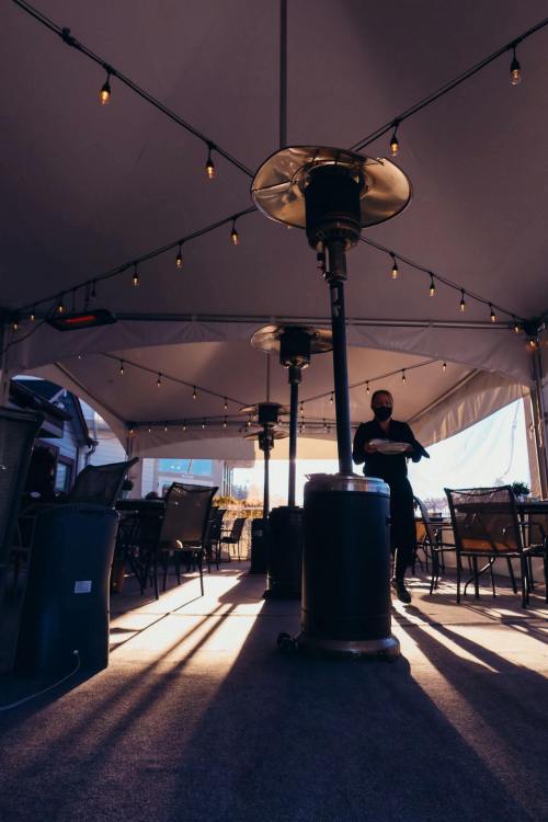 A cozy outdoor dining area with string lights and patio heaters, featuring a person carrying a plate.