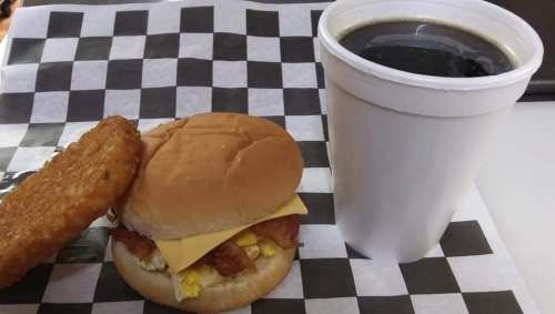 A breakfast sandwich with cheese and bacon, a hash brown, and a cup of black coffee on a checkered paper.