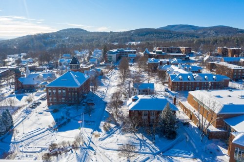 Aerial view of a snowy campus with buildings and trees, surrounded by hills under a clear blue sky.