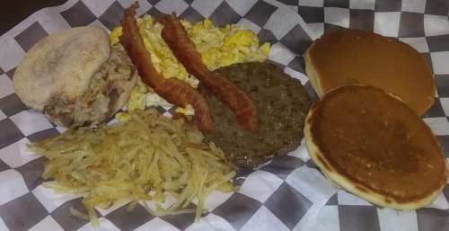 A breakfast plate featuring an English muffin sandwich, scrambled eggs, bacon, sausage patty, hash browns, and pancakes.