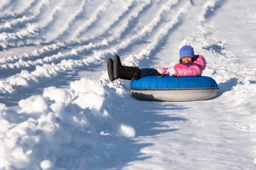 A child in a pink jacket and blue hat smiles while sitting on a blue snow tube in a snowy landscape.