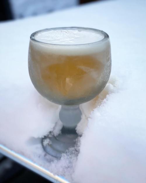 A frosty glass of beer sits on a snowy surface, with ice crystals forming on the glass.