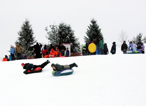 Two children sledding down a snowy hill, surrounded by people enjoying winter activities in the background.