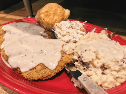 A red plate with two pieces of fried chicken smothered in gravy, mashed potatoes, coleslaw, and a biscuit.