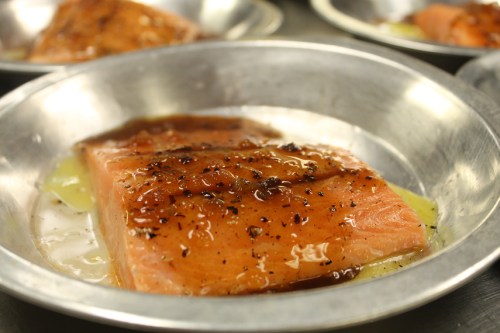 A piece of glazed salmon on a metal plate, garnished with spices and a shiny sauce.