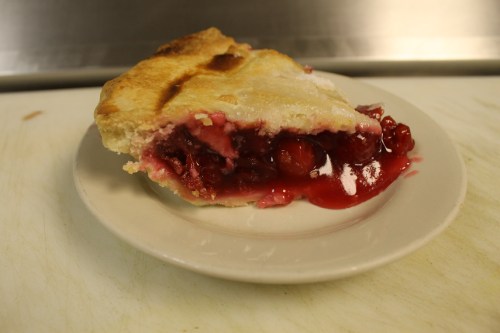 A slice of cherry pie on a white plate, showcasing a flaky crust and vibrant red cherry filling.
