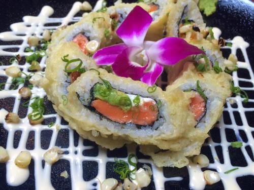 A beautifully plated sushi roll topped with a purple flower, drizzled with sauce and garnished with green onions.