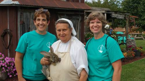 Three women in matching teal shirts pose together outside a rustic building, one holding a rabbit. Colorful flowers are nearby.