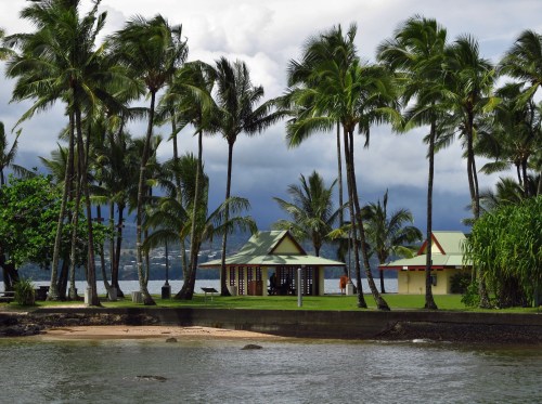 A tropical scene featuring palm trees, a sandy shore, and colorful beach huts by the water under a cloudy sky.