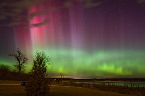 Vibrant auroras in shades of green and pink dance across a night sky, reflecting over a calm body of water.