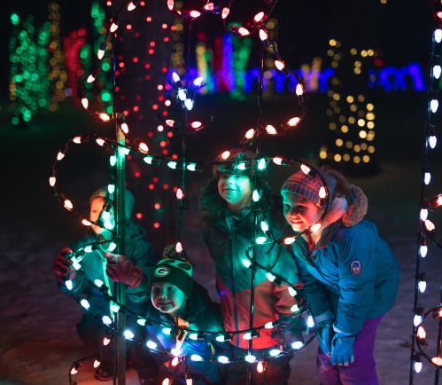 Four children play among colorful holiday lights, smiling and posing in a festive outdoor setting.