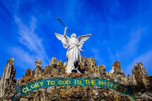 A white angel statue with a trumpet stands atop rocky terrain against a blue sky, with the text "Glory to God in the Highest."