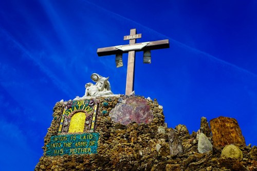 A large cross atop a stone structure with a statue of Mary and Jesus, set against a bright blue sky.