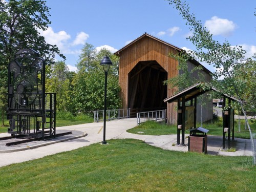 A wooden covered bridge stands beside a pathway, with green trees and a lamp post nearby on a sunny day.