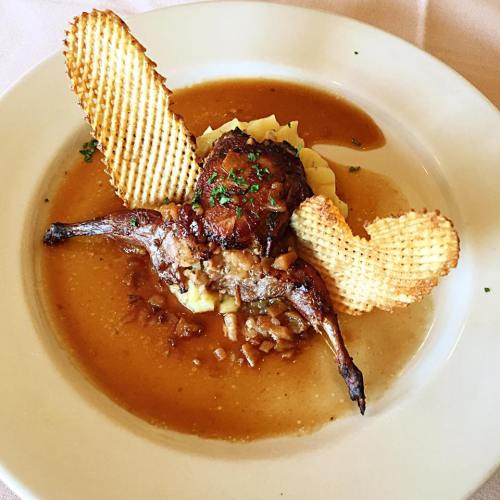 A plated dish featuring a piece of meat on mashed potatoes, garnished with sauce and decorative waffle chips.