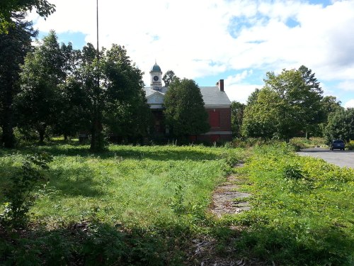 Abandoned building surrounded by overgrown grass and trees, with a clock tower visible in the background under a cloudy sky.