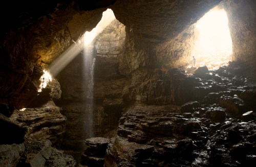 A person stands in a cave illuminated by sunlight, with a waterfall cascading down rocky walls.