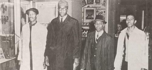 Four men in formal attire walking together in a vintage setting, showcasing a moment from the past.