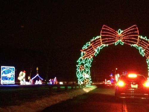 Colorful holiday lights decorate a roadside display, featuring a large bow and festive scenes, viewed from a car at night.