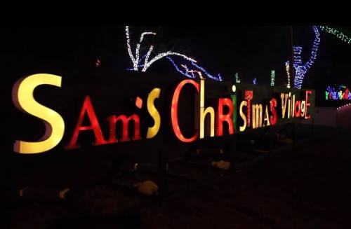 Colorful illuminated sign reading "Sam's Christmas Village" with festive lights in the background.
