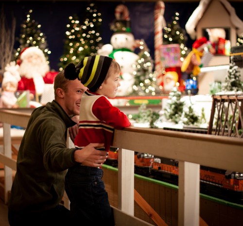 A man and a child watch a festive train display, surrounded by holiday decorations and lights.