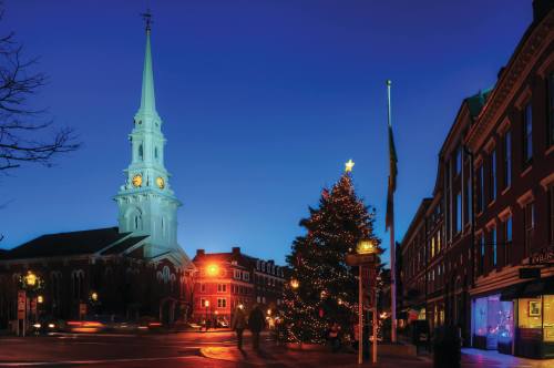 A festive scene featuring a church steeple, a decorated Christmas tree, and buildings illuminated at dusk.