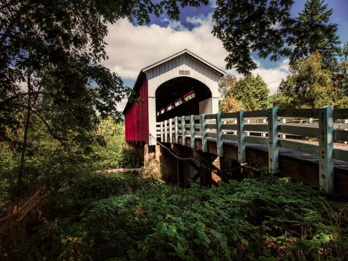 A red and white covered bridge surrounded by lush greenery and trees under a partly cloudy sky.