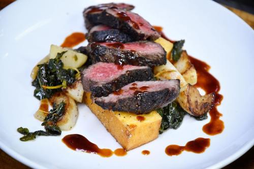 Sliced steak on a bed of polenta, garnished with greens and drizzled with sauce, served on a white plate.