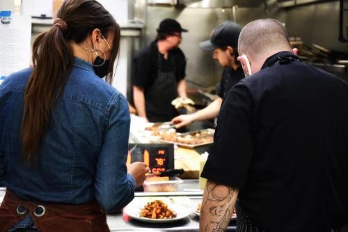 A woman in a denim shirt observes chefs preparing food in a busy kitchen, with a plate of food in the foreground.