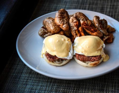 Two eggs Benedict on English muffins with sausage, topped with hollandaise sauce, served with crispy potatoes.