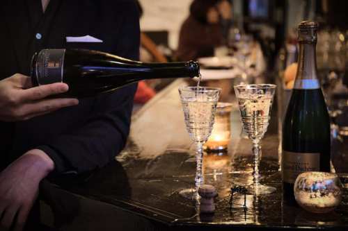 A person pours champagne into two elegant glasses at a bar, with a bottle and candles in the background.