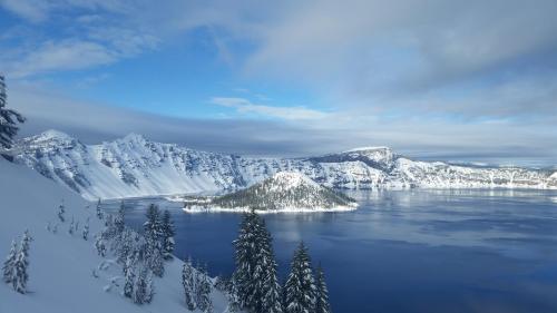 A snowy landscape of Crater Lake, with a calm blue lake surrounded by snow-covered mountains and trees under a cloudy sky.