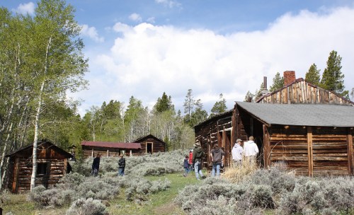 A group of people exploring old wooden cabins surrounded by greenery and trees under a partly cloudy sky.
