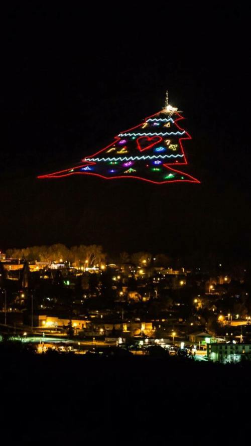 A brightly lit Christmas tree outline on a hillside, glowing against a dark night sky above a city.