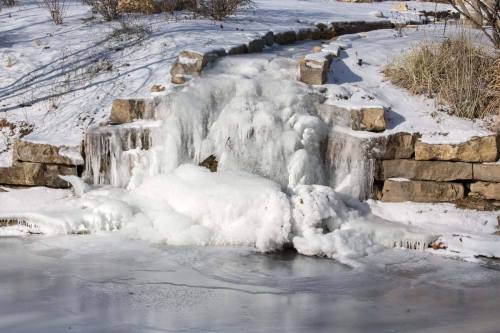 A frozen waterfall cascading over stone steps, surrounded by snow and ice, with a partially frozen pond below.