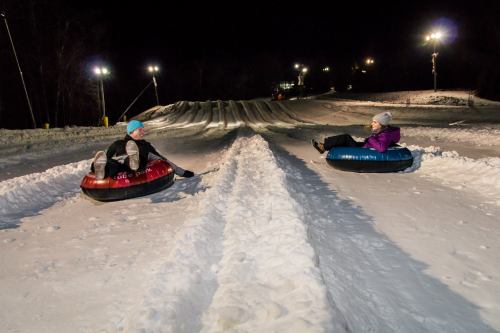 Two people sit in snow tubes on a snowy slope at night, ready to slide down a hill illuminated by lights.