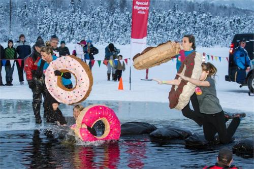 Participants in winter attire jump into a frozen lake, holding large inflatable donuts, during a charity event.