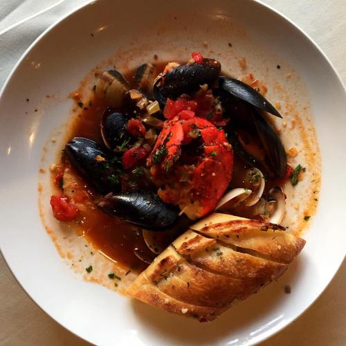 A bowl of seafood pasta with clams, mussels, and a lobster tail, served with a slice of toasted bread.
