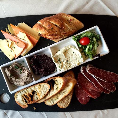 A charcuterie board featuring assorted cheeses, meats, spreads, bread, and a small salad on a black serving platter.