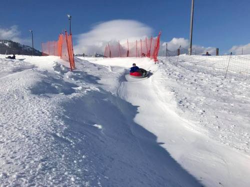A person sleds down a snowy hill, surrounded by orange safety fencing and a clear blue sky.