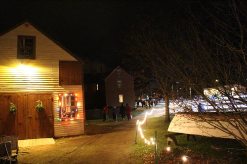A festive night scene with decorated buildings, a pathway lit by lights, and people walking in a charming outdoor setting.